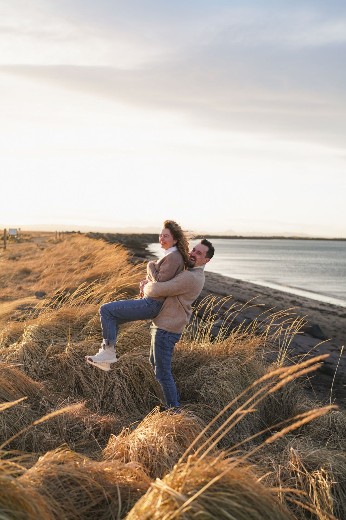 Austin lifting Ana, both laughing, on the shoreline grass at golden hour